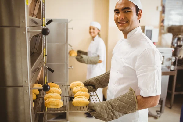 Bäcker lächelt in die Kamera und holt Brötchen aus dem Ofen — Stockfoto