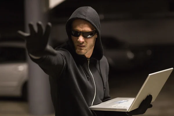 Хакер в балаклаве жестом и с помощью ноутбука — стоковое фото