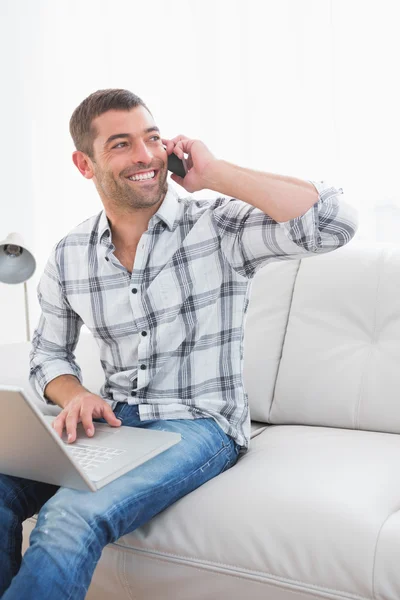 Evde bir dizüstü bilgisayar ile bir cep telefonu üzerinde gülümseyen adam — Stok fotoğraf