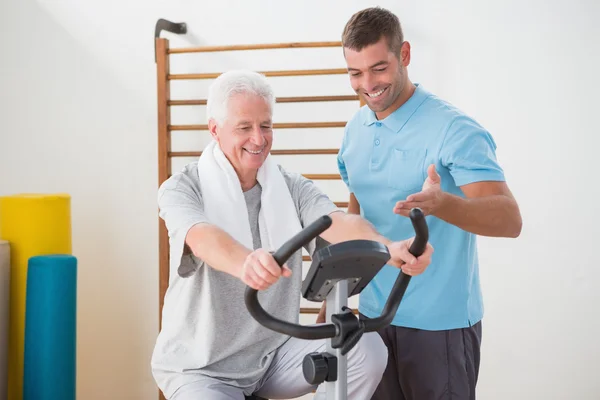 Senior man doing exercise bike with his trainer — Stock Photo, Image