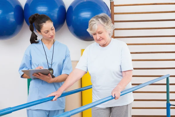 Therapist looking senior woman walking with parallel bars — Stock Photo, Image