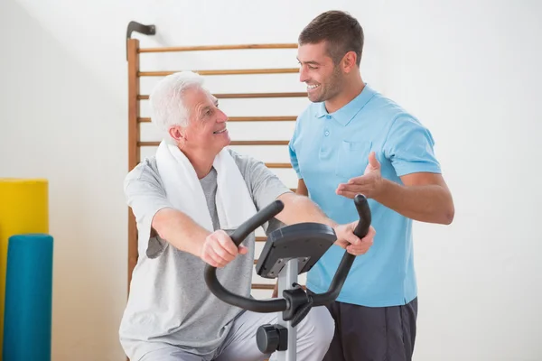 Senior man doen hometrainer met zijn trainer — Stockfoto