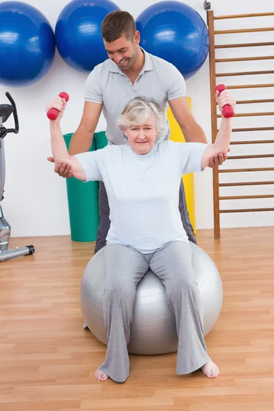 Trainer helpen senior vrouw lift halters op oefening bal — Stockfoto
