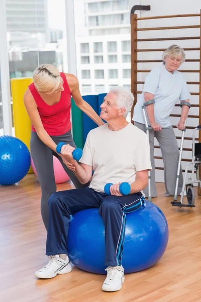 Trainer assisting senior man in lifting dumbbells — Stock Photo, Image
