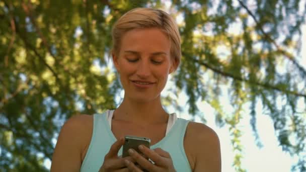 Pasuje blondynka wysyłanie tekstu w parku — Wideo stockowe