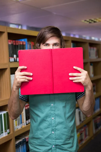 Student covering face with book in library — Stock Photo, Image