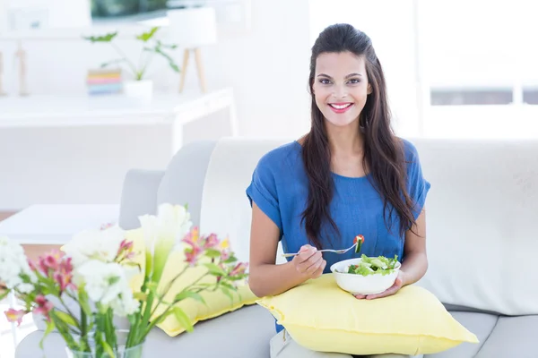 Smiling beautiful brunette sitting on the couch and eating salad — Stock Photo, Image
