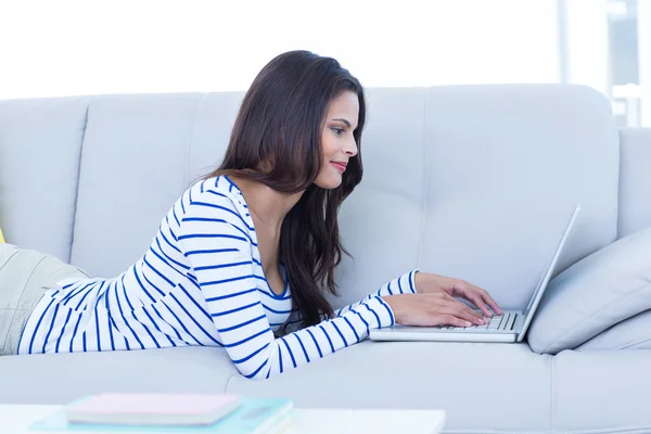 Sonriendo hermosa morena relajándose en el sofá y usando su l —  Fotos de Stock