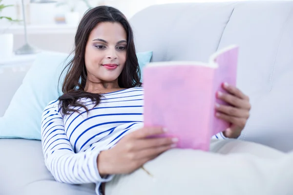 Sorrindo bela morena relaxante no sofá e lendo um b — Fotografia de Stock