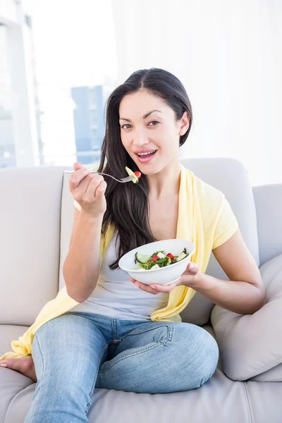 Bonita morena mirando a la cámara y comiendo ensalada en el sofá — Foto de Stock