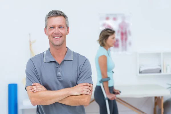 Doctor smiling at camera while his patient standing with crutch — Stock Photo, Image