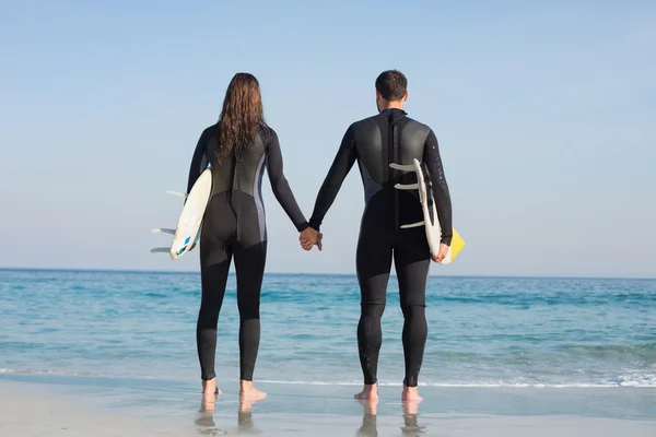 Casal em fatos de mergulho com prancha de surf na praia — Fotografia de Stock