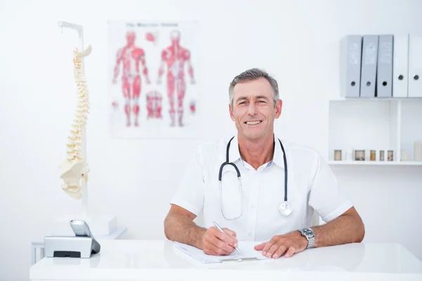 Sonriente doctor escribiendo en el portapapeles en su escritorio — Foto de Stock