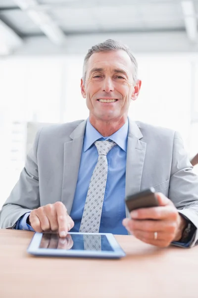 Empresario sonriendo a la cámara con su tableta y su teléfono —  Fotos de Stock