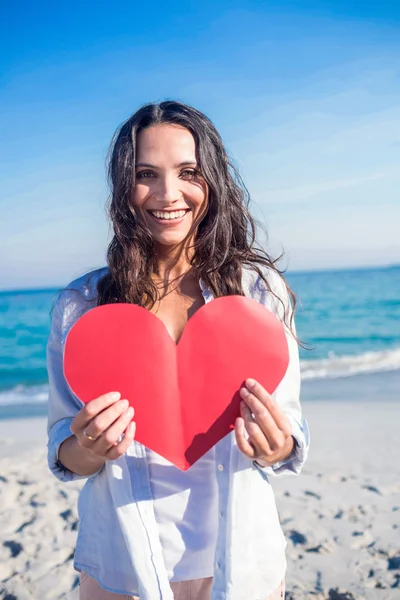 Smiling woman holding heart card at the beach — ストック写真