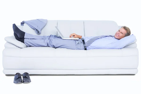 Businessman lying on sofa using his laptop smiling at camera — Stock Photo, Image