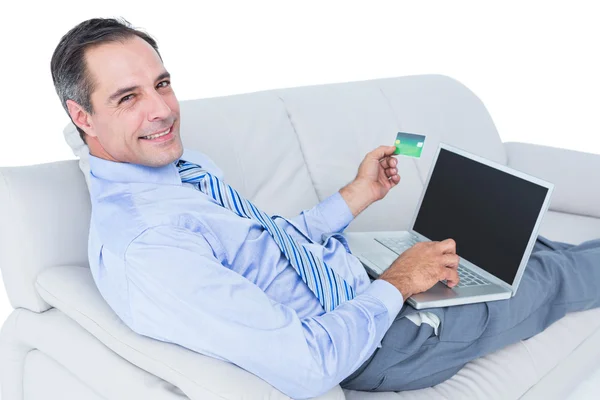 Smiling businessman lying on a sofa holding a card and laptop — Stock Photo, Image