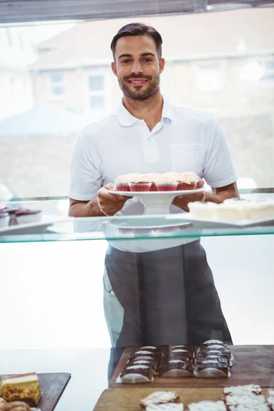 Trabalhador sorrindo segurando cupcakes atrás do balcão — Fotografia de Stock