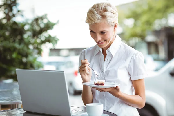 Mujer joven que tiene un pedazo de pastel usando el ordenador portátil —  Fotos de Stock