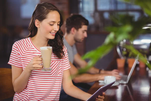 S úsměvem mladá žena líbí latte a použití tabletového počítače — Stock fotografie