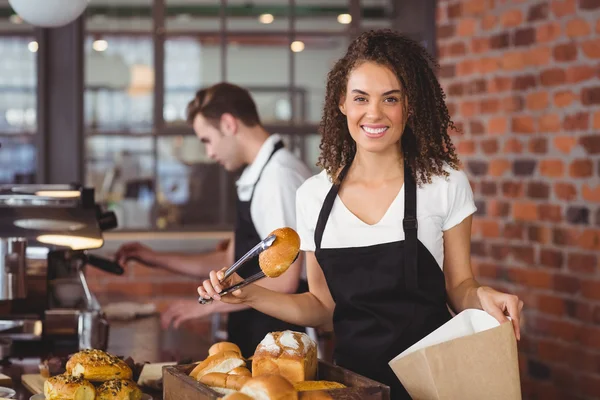 Smiling waitress putting bread roll in paper bag — Stock Photo, Image