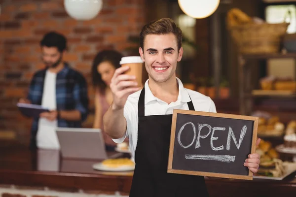 Smiling barista holding take-away cup and open sign