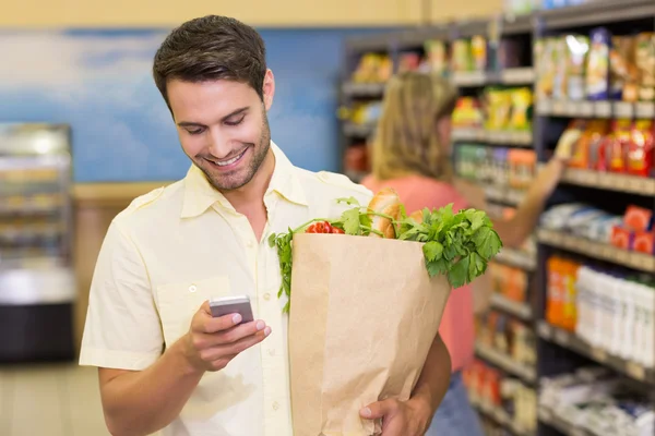 Sonriente hombre guapo comprando productos alimenticios y usando su smartpho —  Fotos de Stock