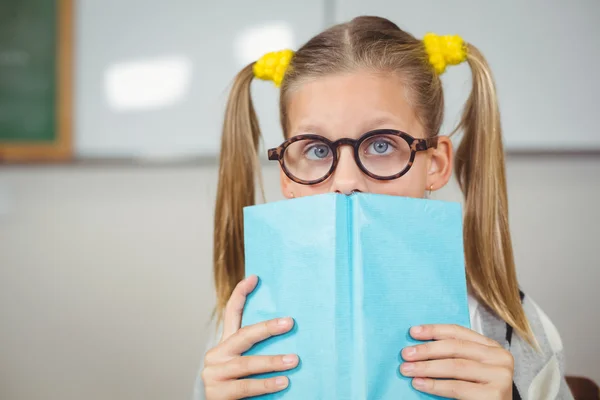 Netter Schüler, der im Klassenzimmer sein Gesicht mit einem Buch bedeckt — Stockfoto