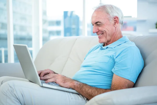 Homem sênior sentado no sofá enquanto usando laptop — Fotografia de Stock