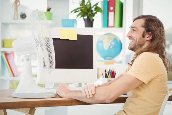 Happy hipster sitting by electric fan on computer desk — Stock Photo, Image