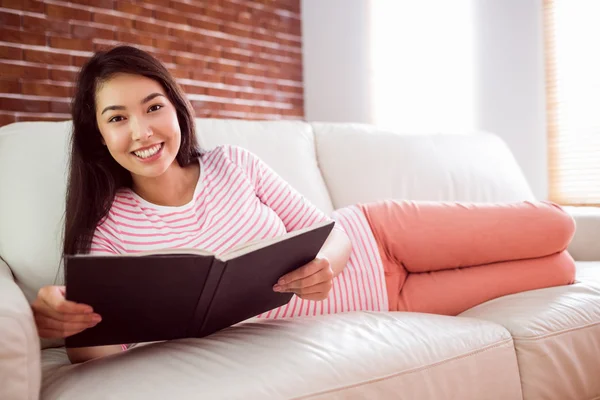 Χαμογελώντας ασιατική γυναίκα στον καναπέ ανάγνωση — Φωτογραφία Αρχείου