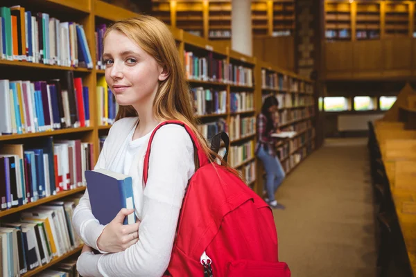 Kitaplıkta bir kitap tutan sırt çantası ile gülümseyen öğrenci — Stok fotoğraf