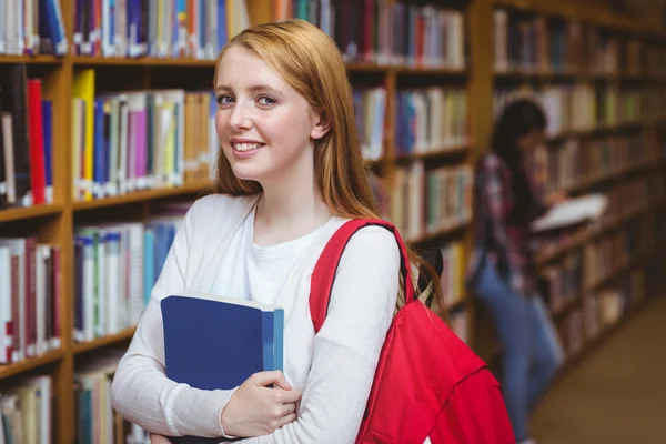 Kitaplıkta bir kitap tutan sırt çantası ile gülümseyen öğrenci — Stok fotoğraf