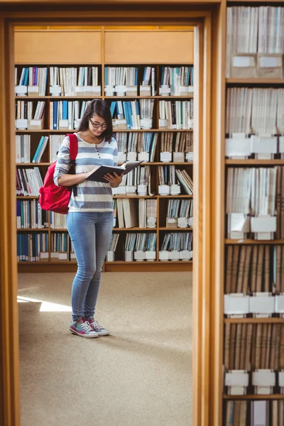 Красивая студентка с рюкзаком читает книгу в библиотеке — стоковое фото