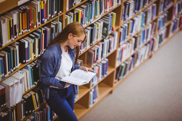 Студент читає книгу в бібліотеці, спираючись на книжкові полиці — стокове фото