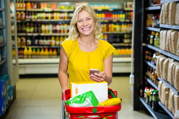 Donna sorridente con smartphone spingendo carrello in corridoio — Foto Stock