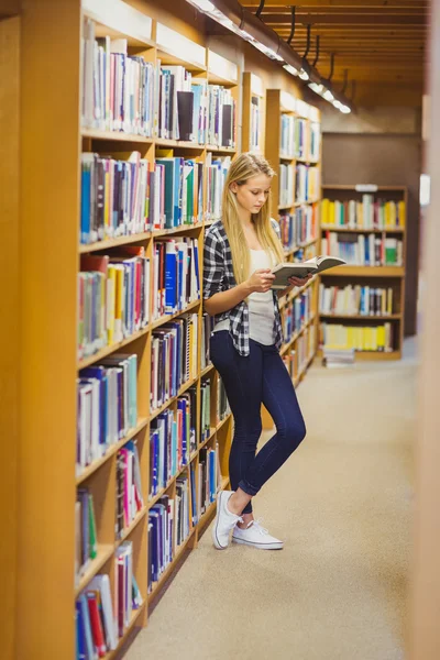 Student reading book next to bookshelf — ストック写真