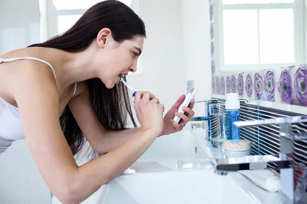 Brunette met behulp van de smartphone tijdens het tandenpoetsen — Stockfoto