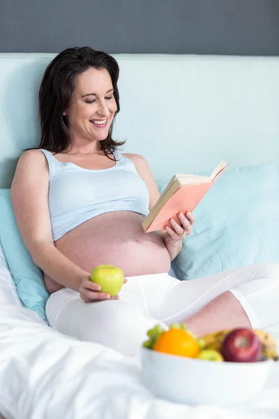 Mujer embarazada comiendo y leyendo libro — Foto de Stock