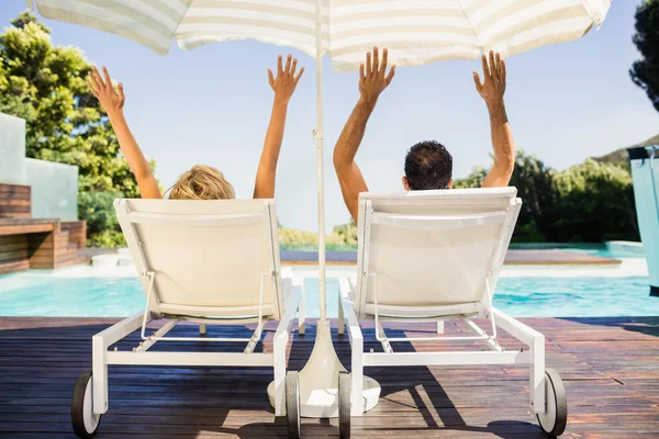 Rear view of couple raising hands and lying on deck chairs — Stock Photo, Image