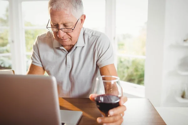 Focalizzato uomo anziano utilizzando computer portatile e bere vino — Foto Stock