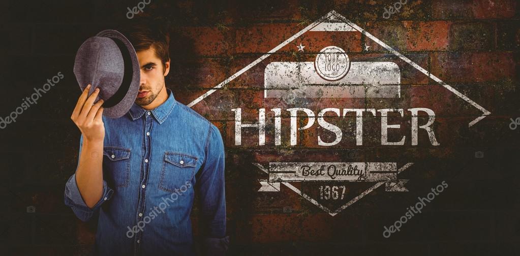 Portrait of hipster holding hat in front of face against texture of bricks wall