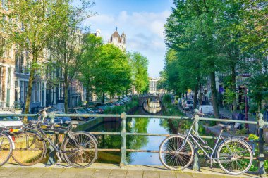 Tarihi binalar, köprüler, köprüler ve ağaçlarla dolu manzaralı köprü raylı bisikletler arka plandaki suya yansıyor, Amsterdam, Hollanda - 19 - 19 Haziran - 2024