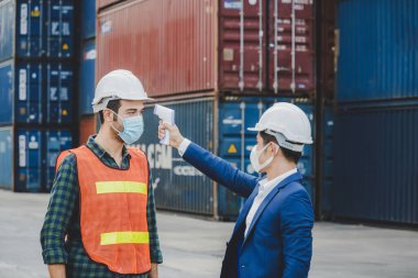 Fabrika işçisi vücut ısısını dijital termometreyle kontrol ediyor. Kargo limanı, endüstriyel, lojistik ithalat ve ihracat konseptinde arka planda kargo konteynırı olan mühendis üzerinde.