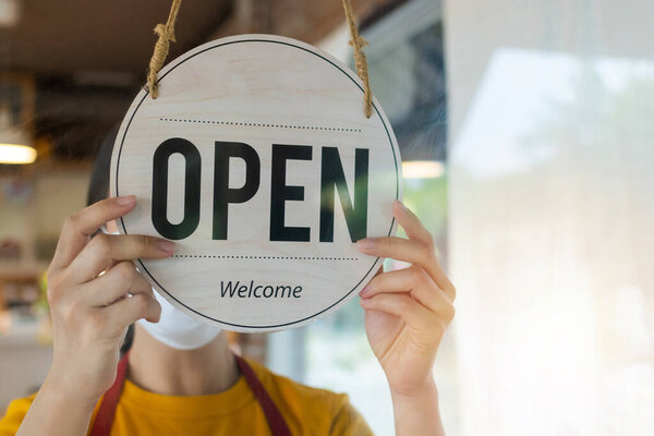 Open. asian small business owner woman wearing protection face mask turning open sign board on glass door for reopening cafe restaurant after coronavirus (covid-19) quarantine. food and drink concept