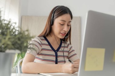 Güzel Asyalı kız öğrenci evde kulaklık ve dizüstü bilgisayarla, ağ iletişimiyle, çevrimiçi toplantı, video konferansı, internet teknolojisi ve çevrimiçi eğitim konseptiyle çevrimiçi çalışıyor.