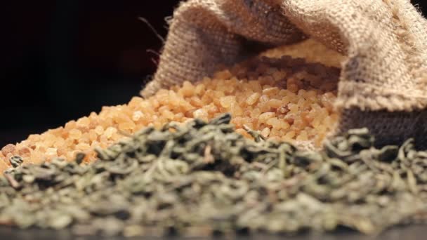 Bruine suiker in gunny zak en een stapel van gedroogde groene thee bladeren, roteren — Stockvideo