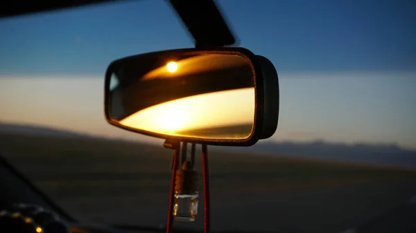 Arabanın dikiz aynası ve şafak. Tepelerin üzerinde turuncu şafak. Araba yüksek hızda gidiyor. Yeşil alanlar, çimenler ve çayırlar görülebilir. Arabanın siyah rengi. Direksiyon, kaput, cam.