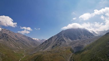 Yüksek dağların tepelerinden bulutlar akar. Rocky uçurumları ve geçitleri. Gökyüzü mavi, güneş ışıl ışıl parlıyor, bazı yerlerde yeşil çimenler büyüyor. Dağ arazisi İHA 'nın yüksekliğinden
