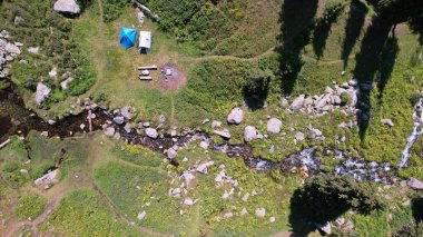Dağ ormanında küçük bir gölet ve bir dere. Nehir yeşil çayırların arasından akar, parlak çiçekler. Kozalaklı ağaçlar büyüyor. Nehir boyunca büyük taşlar. Kamp için bir yer. Birkaç çadır.